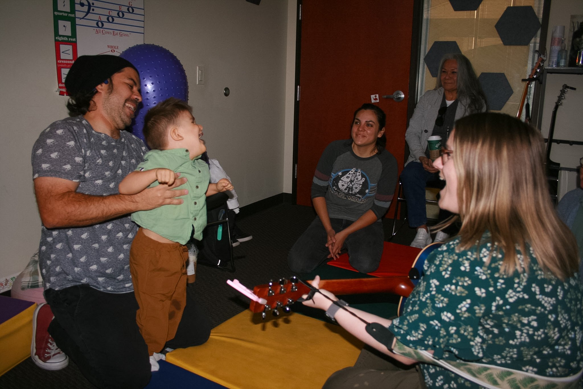 A young child is being held by a man while a woman holding a guitar is smiling at them. Two women are in the background smiling at the scene
