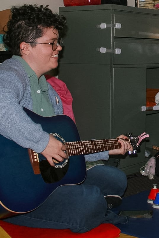 A woman with short brown hair and glasses is holding a guitar while smiling